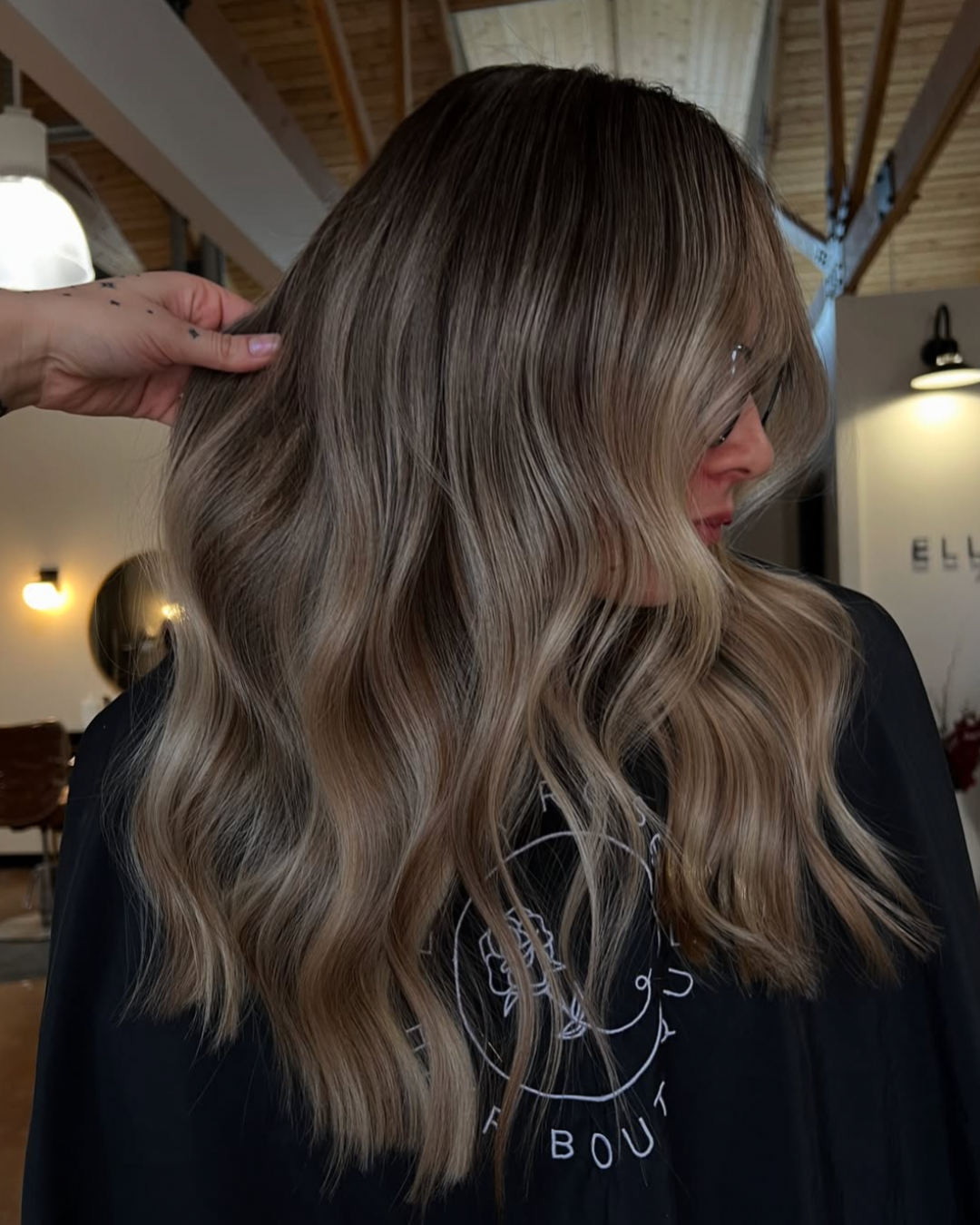 Back view of a woman with long wavy blonde balayage hair featuring blended brunette roots and soft beige blonde highlights.