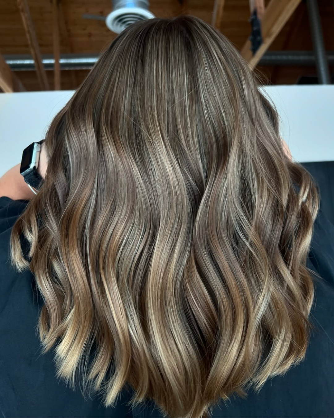 Back view of a woman with shoulder-length lived-in blonde hair featuring darker roots and blended beige blonde tones styled in loose waves.
