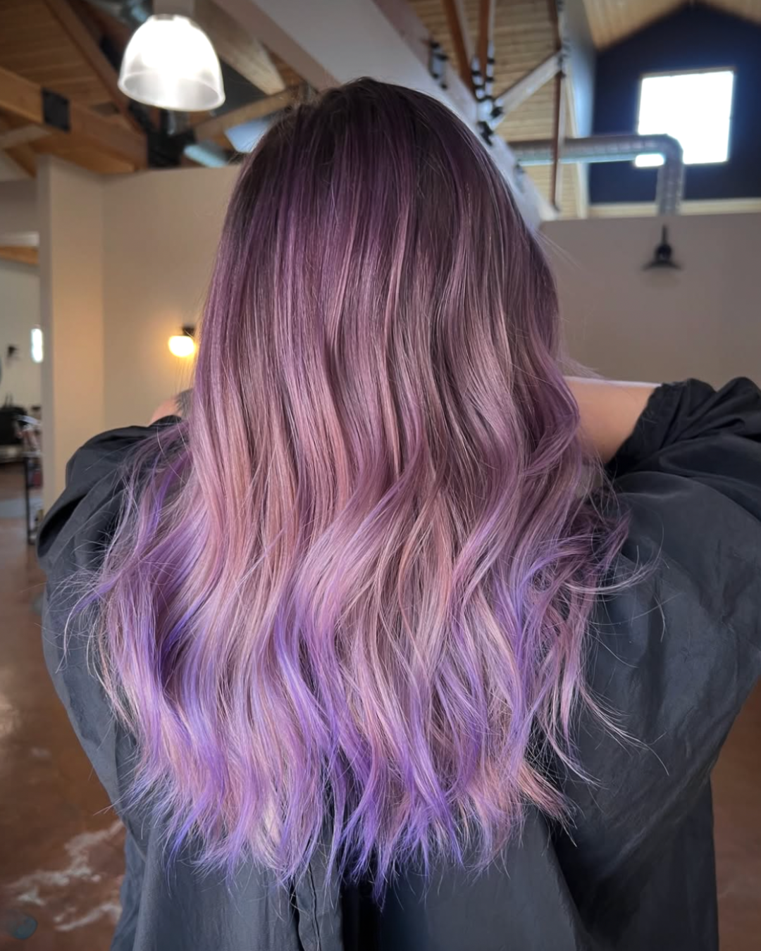 Back view of a woman with long wavy lavender hair featuring soft pastel and deep purple tones throughout.