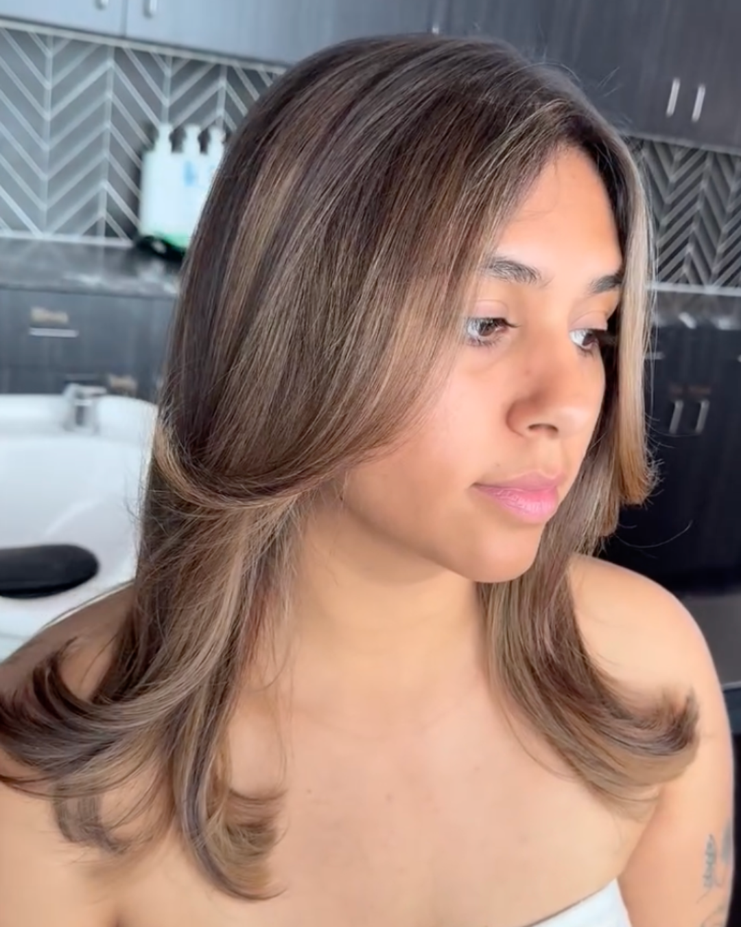 Woman with medium-length layered brunette hair featuring soft caramel highlights and a smooth blowout.