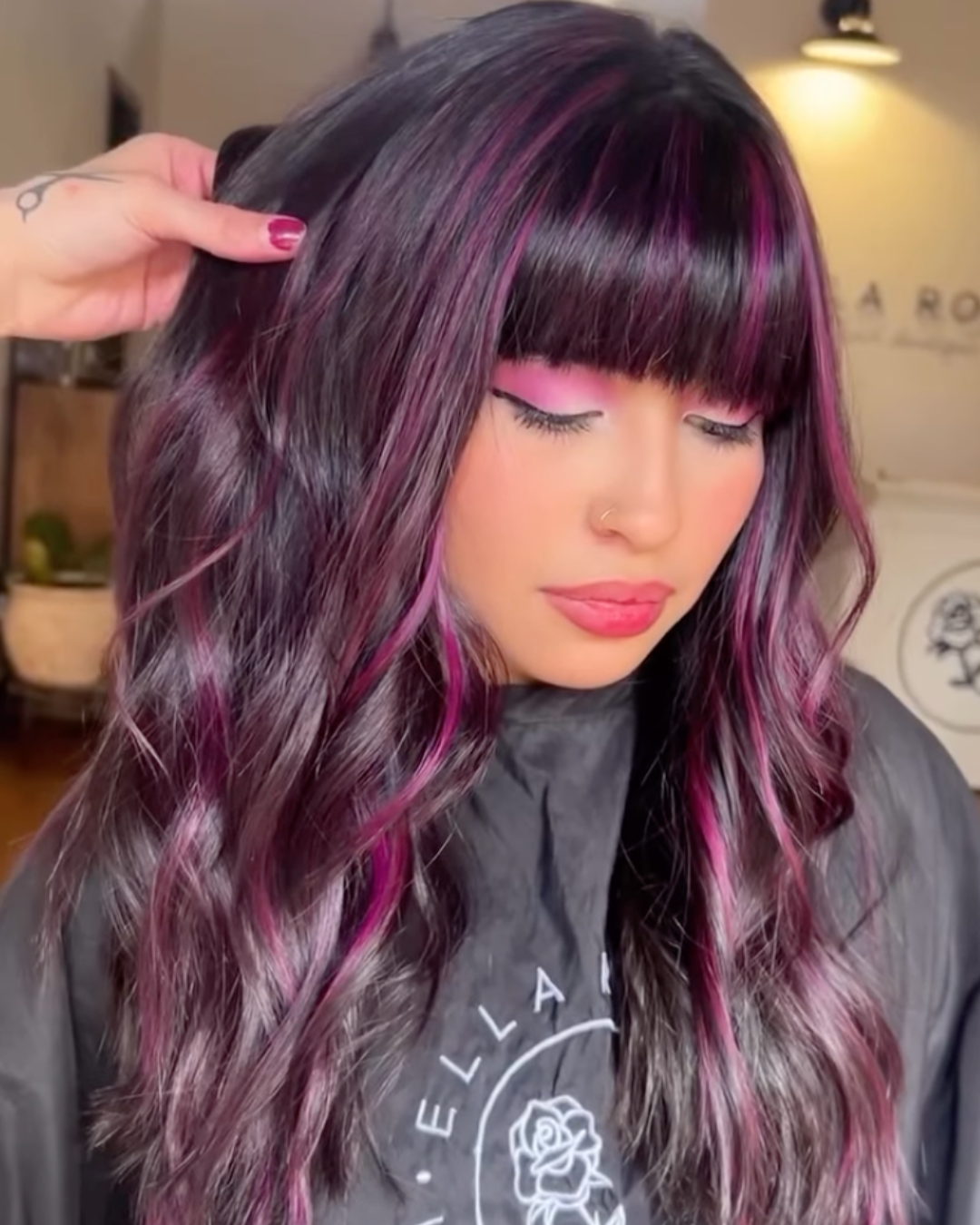 Woman with long wavy dark hair featuring vibrant magenta highlights throughout and full bangs.